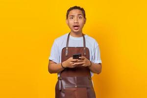 retrato de jovem asiático vestindo avental parecendo surpreso e segurando o smartphone sobre fundo amarelo foto