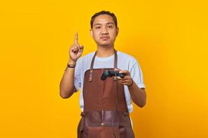 retrato de um jovem asiático bonito usando avental, segurando o controlador de jogo, tendo uma ótima ideia isolada no fundo amarelo foto