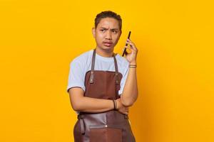 retrato de jovem asiático vestindo avental, parecendo zangado enquanto fala no smartphone sobre fundo amarelo foto