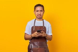 retrato de jovem asiático vestindo avental, parecendo zangado e segurando o smartphone sobre fundo amarelo foto