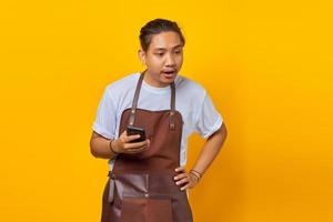 retrato de jovem asiático vestindo avental surpreso ao ver a mensagem recebida no smartphone em fundo amarelo foto