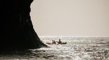 feliz casal caiaques dentro a inflável caiaque em a mar às pôr do sol. casal canoagem dentro a mar perto a ilha com montanhas. pessoas caiaque dentro vida jaquetas velejar. costas Visão foto