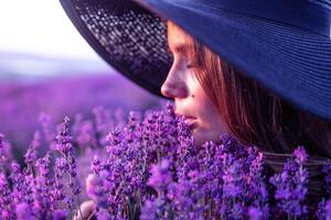 mulher lavanda campo. feliz despreocupado mulher dentro Preto vestir e chapéu com ampla borda cheirando uma florescendo lavanda em pôr do sol. perfeito para inspirado e caloroso conceitos dentro viagem e desejo de viajar. fechar acima foto