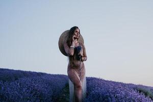 mulher lavanda campo. feliz despreocupado mulher dentro bege vestir e chapéu com ampla borda cheirando uma florescendo lavanda em pôr do sol. perfeito para inspirado e caloroso conceitos dentro viagem e desejo de viajar. fechar acima foto