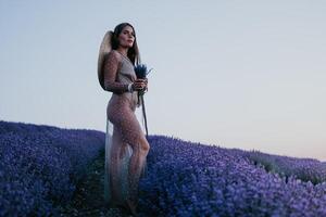 mulher lavanda campo. feliz despreocupado mulher dentro bege vestir e chapéu com ampla borda cheirando uma florescendo lavanda em pôr do sol. perfeito para inspirado e caloroso conceitos dentro viagem e desejo de viajar. fechar acima foto