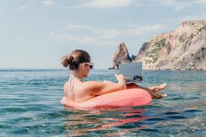 mulher trabalhador autonomo trabalho em computador portátil natação dentro mar em Rosa inflável anel. feliz turista dentro oculos de sol flutuando em inflável rosquinha e trabalhando em computador portátil computador dentro calma oceano. controlo remoto trabalhando qualquer lugar foto