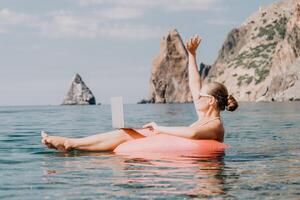 mulher trabalhador autonomo trabalho em computador portátil natação dentro mar em Rosa inflável anel. feliz turista dentro oculos de sol flutuando em inflável rosquinha e trabalhando em computador portátil computador dentro calma oceano. controlo remoto trabalhando qualquer lugar foto