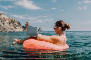 mulher trabalhador autonomo trabalho em computador portátil natação dentro mar em Rosa inflável anel. feliz turista dentro oculos de sol flutuando em inflável rosquinha e trabalhando em computador portátil computador dentro calma oceano. controlo remoto trabalhando qualquer lugar foto