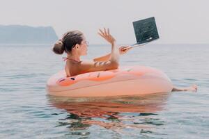 mulher trabalhador autonomo trabalho em computador portátil natação dentro mar em Rosa inflável anel. feliz turista flutuando em inflável rosquinha e trabalhando em computador portátil computador dentro calma oceano. freelance, controlo remoto trabalhando qualquer lugar foto