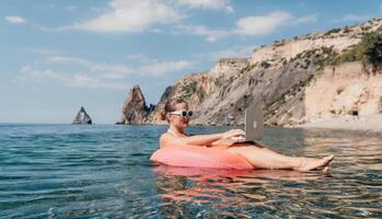 mulher trabalhador autonomo trabalho em computador portátil natação dentro mar em Rosa inflável anel. feliz turista dentro oculos de sol flutuando em inflável rosquinha e trabalhando em computador portátil computador dentro calma oceano. controlo remoto trabalhando qualquer lugar foto