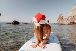 mulher mar e aí. fechar acima retrato do feliz jovem caucasiano mulher com grandes cabelo dentro santa chapéu olhando às Câmera e sorridente. fofa mulher retrato dentro uma branco bikini posando em sup borda dentro a mar foto