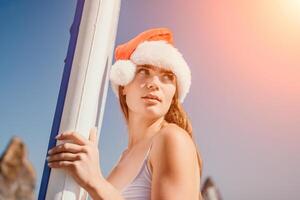 mulher mar e aí. fechar acima retrato do feliz jovem caucasiano mulher com grandes cabelo dentro santa chapéu olhando às Câmera e sorridente. fofa mulher retrato dentro uma branco bikini posando em sup borda dentro a mar foto