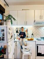 pequeno menina senta em a mesa dentro a cozinha perto a geladeira foto