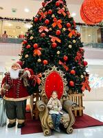 budva, Montenegro - 25 dezembro 2022. pequeno menina senta em a poltrona perto a Natal árvore dentro a Shopping foto