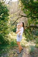 sorridente mãe com pequeno filha em pé perto árvore dentro ensolarado parque foto