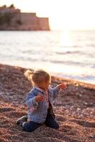 pequeno menina sentado em dela joelhos em uma seixo de praia com dela mãos acima e dedos espalhar foto