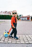 pequeno menina é empurrando uma brinquedo carrinho ao longo a pavimentação pedras, girando dela cabeça para a lado foto