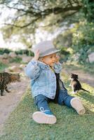 pequeno menina dentro uma chapéu senta em verde Relva perto uma gatinho e uma gato foto