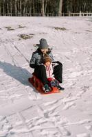 sorridente mãe com uma pequeno criança trenó baixa uma coberto de neve Colina foto