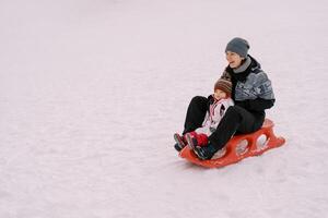 rindo mãe com uma pequeno menina passeio uma trenó em uma Nevado Colina foto