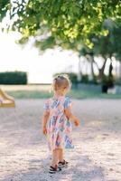 pequeno menina é caminhando em a Parque infantil debaixo uma verde árvore. costas Visão foto