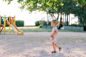 pequeno menina anda em em a Parque infantil olhando às a deslizar. lado Visão foto