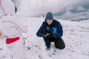 pequeno menina parece às Papai fazer uma pequeno boneco de neve em uma Nevado Colina foto