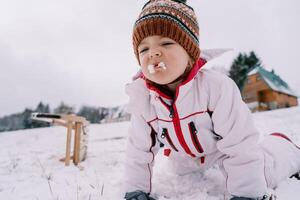 pequeno menina senta em dela joelhos em uma Nevado Colina e tentativas a neve com uma careta foto
