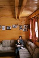 mãe lê uma livro para uma pequeno menina sentado em dela colo em a sofá dentro uma de madeira casa foto