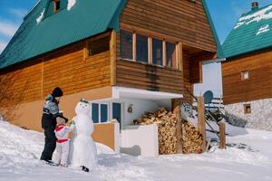 mãe e pequeno menina decorar uma boneco de neve perto uma pilha de lenha do uma de madeira chalé dentro a neve foto