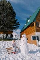 pequeno menina senta em uma trenó perto uma boneco de neve dentro a pátio do uma de madeira chalé foto