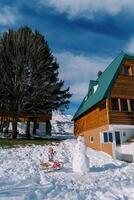 pequeno menina come a partir de uma prato sentado em uma trenó perto uma boneco de neve Próximo para uma de madeira casa foto