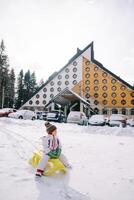 pequeno menina senta em uma trenó dentro a neve perto carros estacionado perto uma triangular hotel foto