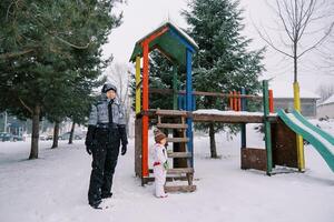 mãe e pequeno menina ficar de pé perto uma coberto de neve colorida deslizar entre a árvores foto