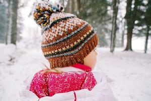 pequeno menina carrinhos debaixo queda de neve dentro uma Nevado floresta e parece para a lado. costas Visão foto