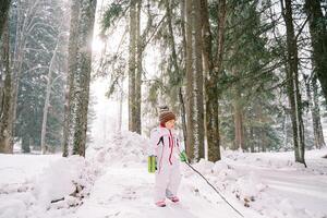 pequeno menina com uma bastão dentro dela mão carrinhos dentro uma Nevado floresta e parece longe foto