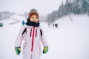 pequeno sorridente menina dentro uma esqui terno carrinhos em uma esqui declive foto