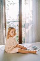 pequeno menina senta em uma cama com uma cenário livro e parece ausente. lado Visão foto