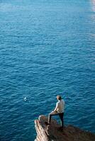 jovem homem carrinhos em uma pedra borda acima a mar e parece para dentro a distância. costas Visão foto