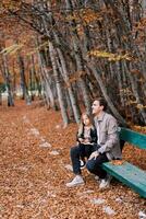 pequeno menina senta em a colo do uma sorridente Papai em uma Banco dentro a outono floresta foto