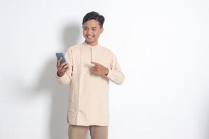 retrato do jovem animado ásia muçulmano homem dentro Koko camisa segurando Móvel telefone, apontando e mostrando produtos com dedo. social meios de comunicação conceito. isolado imagem em branco fundo foto