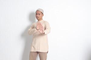 retrato do desagradável ásia muçulmano homem dentro Koko camisa com calota craniana formando uma mão gesto para evitar alguma coisa ou mau coisas. isolado imagem em branco fundo foto