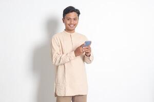 retrato do jovem animado ásia muçulmano homem dentro Koko camisa segurando Móvel telefone com sorridente expressão em face. social meios de comunicação e Internet conceito. isolado imagem em branco fundo foto