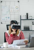 mulher examina papelada enquanto vestindo uma virtual realidade fone de ouvido, sentado às dela escrivaninha com uma computador portátil. foto