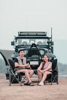 homem e mulher rir alegremente enquanto sentado dentro acampamento cadeiras, com uma fora da estrada carro dentro a pano de fundo, desfrutando seus ao ar livre aventura. foto