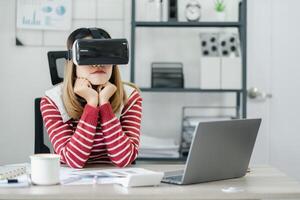 mulher senta às dela escrivaninha com uma virtual realidade fone de ouvido sobre, mãos debaixo queixo, olhando contemplativo. foto