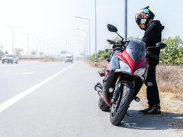 motociclista homem em pé com motocicleta estacionado em lado do estrada foto