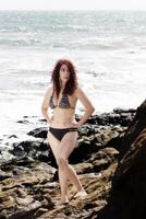latina mulher em pé em pedras dentro bikini com oceano foto