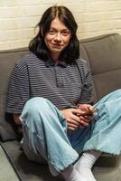 sorridente jovem mulher a partir de geração z usando uma Smartphone em a sofá às casa foto