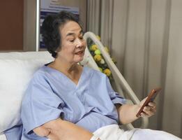 alegre ásia Senior fêmea paciente sentado dentro hospital cama, sorridente e olhando às Móvel telefone, vídeo chamando para dela família. idosos saúde conceito. foto